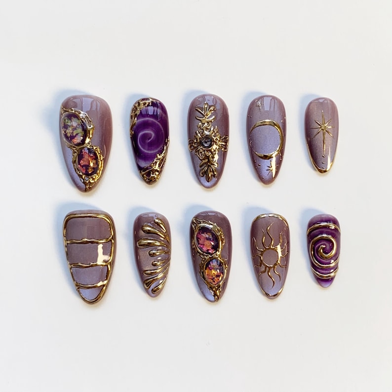Pode incluir: Um conjunto de dez unhas posti&ccedil;as lavanda com enfeites dourados. Os desenhos incluem espirais douradas, pedras preciosas, um sol, uma lua e uma estrela. As unhas s&atilde;o ovais e dispostas sobre uma superf&iacute;cie branca.