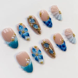 May include: A collection of artificial nails with various designs. Some nails feature a nude base with blue and gold accents, while others showcase intricate blue floral patterns and pearl embellishments. The nails are arranged on a white surface.