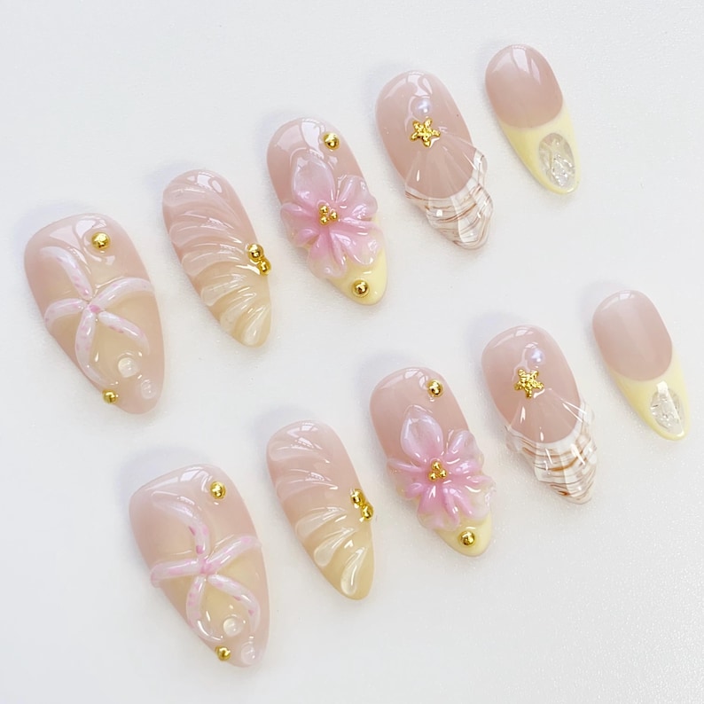Pu&ograve; includere: Un set di dieci unghie finte con vari design. Le unghie presentano una base rosa chiaro con elementi decorativi come borchie dorate, fiori rosa e dettagli chiari e strutturati. Alcune unghie hanno una punta gialla.