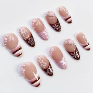 May include: A collection of artificial nails in various designs. The nails feature a nude base with pink, white, and brown accents. Some nails have floral patterns, pearl embellishments, and star-shaped decorations.