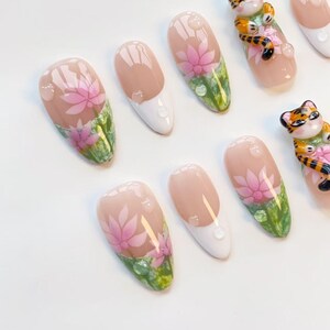 May include: A set of artificial nails with a floral design. The nails feature a pink lotus flower design over a green and white base. Some nails have a clear water droplet design. Two nails have a tiger design.