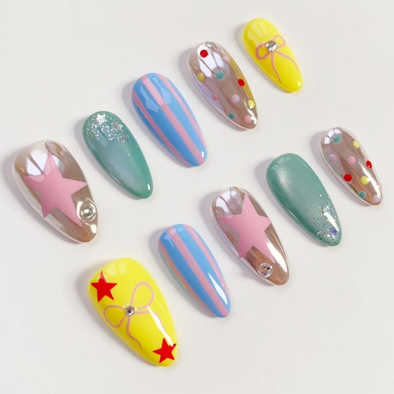 K&ouml;nnte beinhalten: Eine Sammlung k&uuml;nstlicher N&auml;gel in verschiedenen Designs. Die N&auml;gel weisen eine Reihe von Farben auf, darunter Gelb, Blau, Rosa und Gr&uuml;n. Einige N&auml;gel haben Stern-, Streifen- und Punktmuster, w&auml;hrend andere Glitzer- oder Schleifenakzente aufweisen. Jeder Nagel hat ein gl&auml;nzendes Finish.