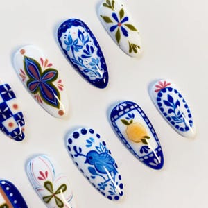 May include: A set of almond-shaped press-on nails featuring hand-painted designs in blue, white, yellow, and green. Patterns include floral motifs, a blue bird, a yellow lemon, geometric checkered squares, and intricate botanical scrollwork.