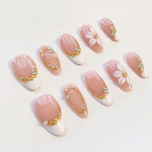 Puede incluir: Un juego de diez u&ntilde;as postizas con base color nude y puntas blancas. Los dise&ntilde;os incluyen detalles de filigrana dorada, peque&ntilde;os diamantes de imitaci&oacute;n transparentes y adornos florales blancos en relieve en dos de las u&ntilde;as almendradas.