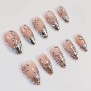May include: Set of ten almond-shaped artificial nails. Each nail features a nude-colored base with a silver, ornate, swirling design and a silver metallic tip. The nails are arranged on a white surface.