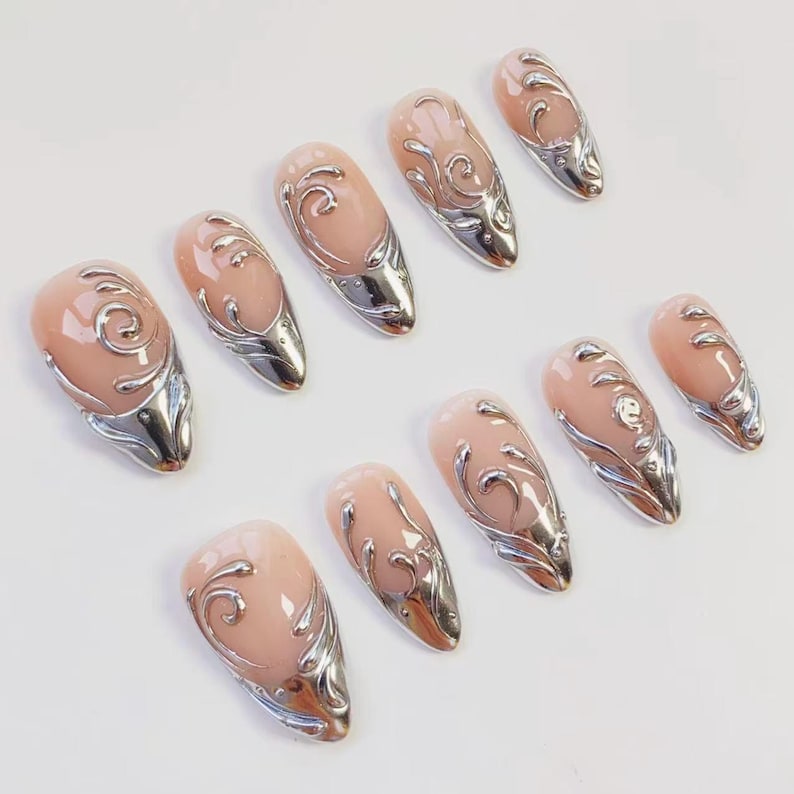 May include: Set of ten almond-shaped artificial nails. Each nail features a nude-colored base with a silver, ornate, swirling design and a silver metallic tip. The nails are arranged on a white surface.