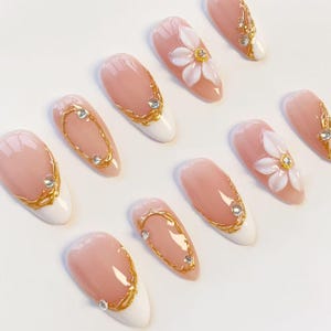 Puede incluir: Un juego de u&ntilde;as postizas en forma de almendra con base color nude y puntas blancas. Los dise&ntilde;os incluyen detalles de filigrana dorada en relieve, peque&ntilde;os diamantes de imitaci&oacute;n transparentes y p&eacute;talos de flores blancos en 3D con centros dorados.