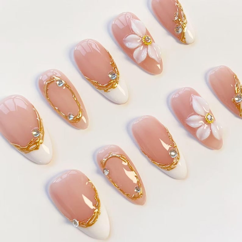 Puede incluir: Un juego de u&ntilde;as postizas en forma de almendra con base color nude y puntas blancas. Los dise&ntilde;os incluyen detalles de filigrana dorada en relieve, peque&ntilde;os diamantes de imitaci&oacute;n transparentes y p&eacute;talos de flores blancos en 3D con centros dorados.