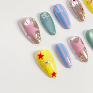 K&ouml;nnte beinhalten: Eine Sammlung k&uuml;nstlicher Fingern&auml;gel in verschiedenen Designs. Einige N&auml;gel sind transparent mit rosa Sternen, andere sind t&uuml;rkis mit Glitzer, und einige haben blau-rosa Streifen. Ein Nagel ist gelb mit roten Sternen und einer Schleife.