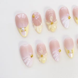 May include: A collection of artificial nails in shades of light pink and yellow. Some nails are decorated with white floral patterns, gold details, and tiny pearl accents. The nails are oval-shaped.