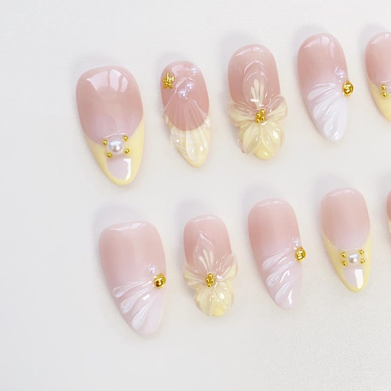 May include: A collection of artificial nails in shades of light pink and yellow. Some nails are decorated with white floral patterns, gold details, and tiny pearl accents. The nails are oval-shaped.
