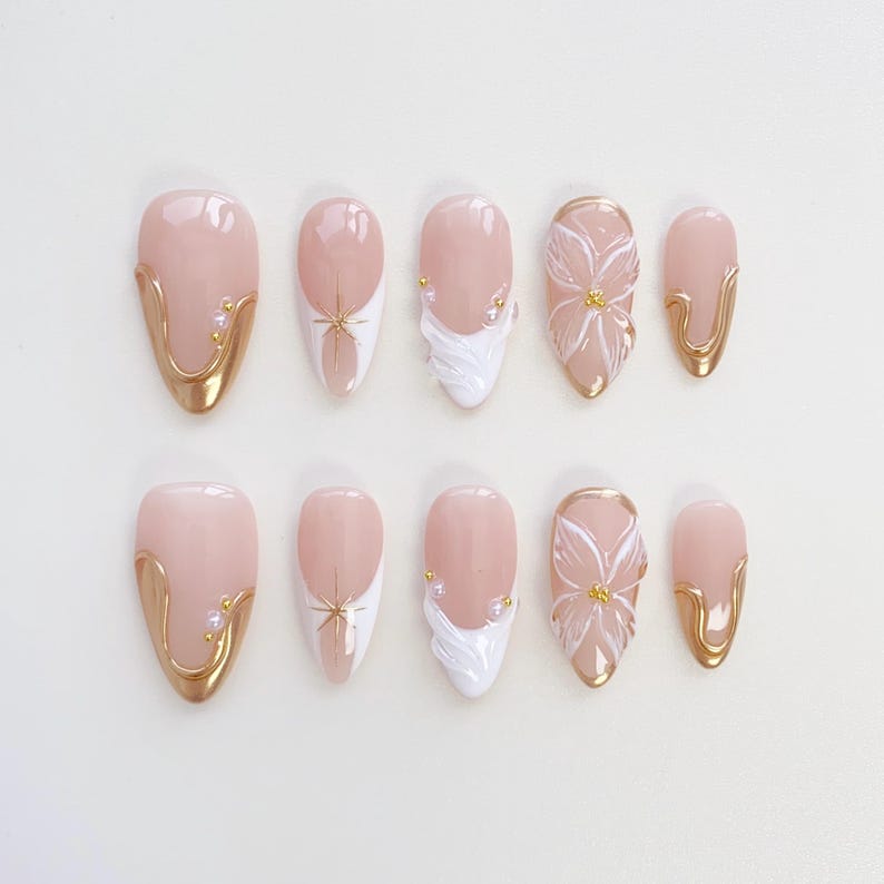 May include: A set of ten almond-shaped false nails. The nails have a pale pink base with gold accents, white floral designs, and small pearl embellishments. Some nails have a gold border.