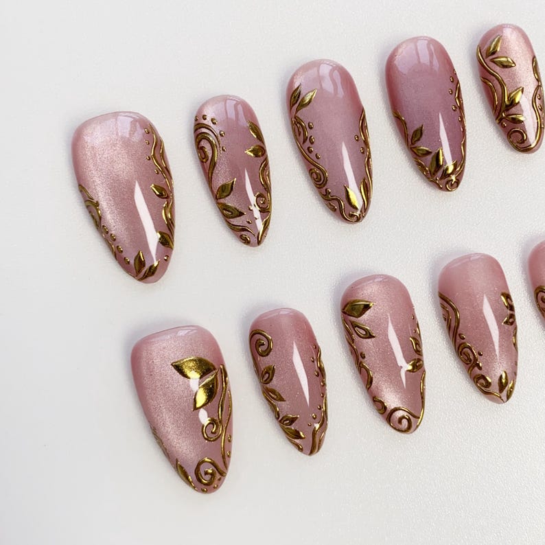 May include: A set of press-on nails in a soft pink hue, adorned with intricate gold leaf and swirl designs. The nails have a glossy finish and are oval-shaped, offering a touch of elegance and sophistication. The gold accents create a striking contrast against the pink.