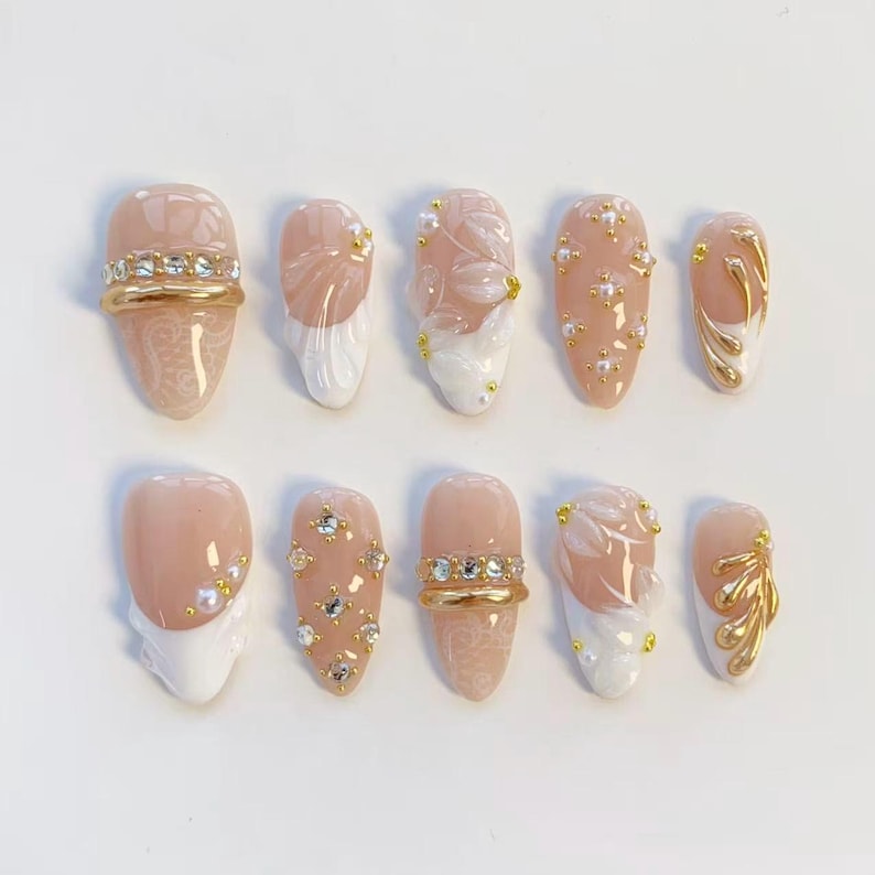 May include: A set of ten artificial nails in various designs. The nails feature a light pink base with white tips, gold accents, and rhinestones. Some nails have floral patterns, while others have gold bands with rhinestones.