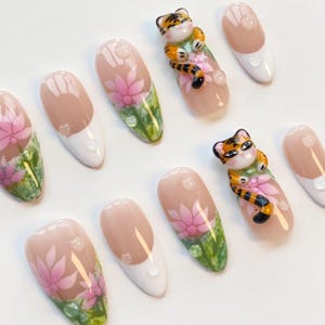 May include: A set of almond-shaped false nails with a light pink base, white tips, and a floral design. Some nails feature a 3D tiger figurine. The nails have a glossy finish and water droplet accents.