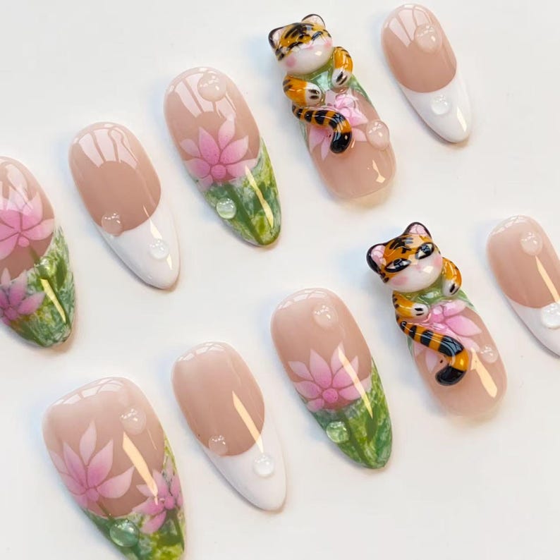 May include: A set of almond-shaped false nails with a light pink base, white tips, and a floral design. Some nails feature a 3D tiger figurine. The nails have a glossy finish and water droplet accents.