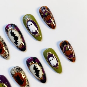 May include: Halloween-themed nail art featuring various designs. Some nails have a white ghost, a black bat, or a pumpkin in a gold frame. The nails are painted in shades of green, purple, and orange.