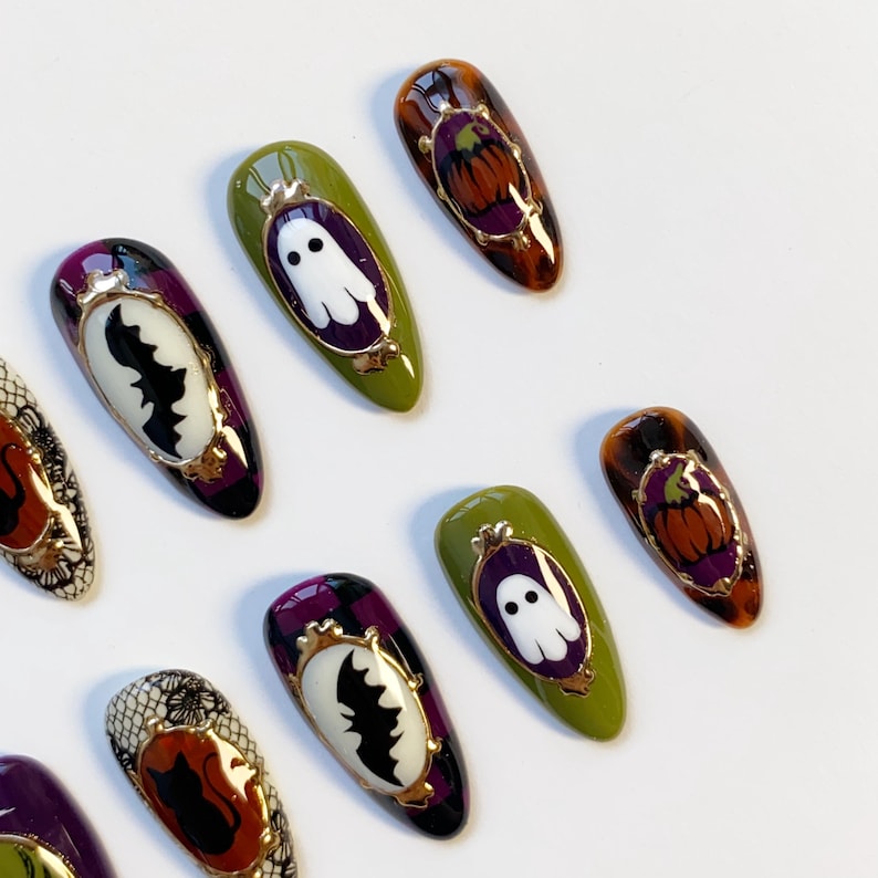 May include: Halloween-themed nail art featuring various designs. Some nails have a white ghost, a black bat, or a pumpkin in a gold frame. The nails are painted in shades of green, purple, and orange.