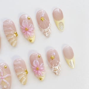 Pu&ograve; includere: Un set di unghie finte a forma di mandorla. Le unghie sono principalmente rosa chiaro con punte gialle, decorate con borchie dorate, fiori rosa 3D e dettagli a stella e perla. Alcune unghie hanno un motivo a quadri.