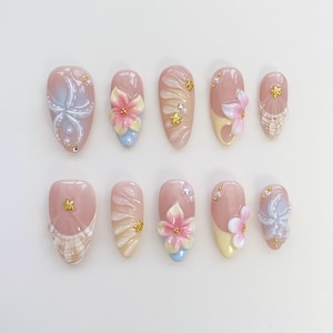 May include: A set of ten artificial nails in various designs. Each nail is a light pink color with different embellishments, including 3D flowers, pearls, and gold accents. Some nails feature a seashell design or a gradient effect.