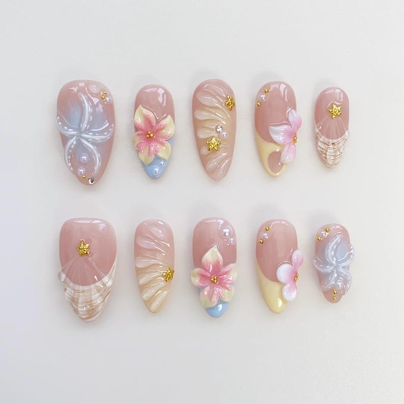 May include: A set of ten artificial nails in various designs. Each nail is a light pink color with different embellishments, including 3D flowers, pearls, and gold accents. Some nails feature a seashell design or a gradient effect.
