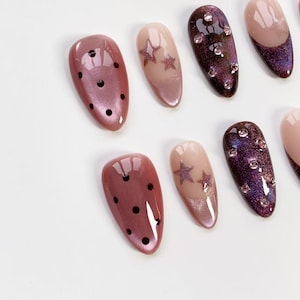 May include: A collection of artificial nails in various designs. Some nails are a shimmery mauve with black polka dots. Others are a gradient of nude to mauve with star accents. Several nails are a deep purple with clear gem embellishments.
