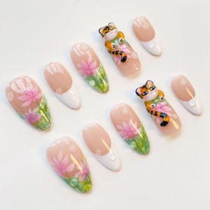 May include: A set of ten artificial nails with various designs. Some nails feature a pink lotus flower design with green leaves and water droplets. Other nails have a white tip or a small tiger figurine.