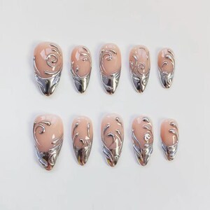 May include: Set of ten artificial nails with a light pink base and silver metallic accents. Each nail features a silver swirl design and a silver metallic tip. The nails are oval-shaped.