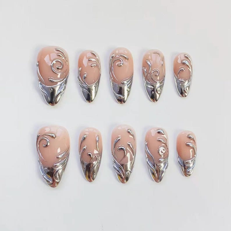 May include: Set of ten artificial nails with a light pink base and silver metallic accents. Each nail features a silver swirl design and a silver metallic tip. The nails are oval-shaped.