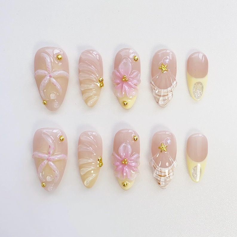 Pu&ograve; includere: Un set di dieci unghie finte con vari design. Le unghie presentano una base rosa chiaro con elementi decorativi come fiori, spirali e accenti dorati. Alcune unghie hanno una punta gialla con glitter argentati.