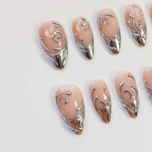 May include: A set of artificial nails with a light pink base and silver metallic accents. Each nail features a swirling design and a silver metallic tip. The nails are oval-shaped and arranged on a white surface.