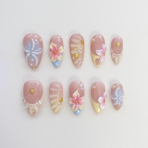 May include: A set of ten artificial nails in various designs. Each nail is a light pink color with detailed floral and abstract designs. Some nails feature 3D flowers in pink, yellow, and blue, while others have gold accents and pearl embellishments.