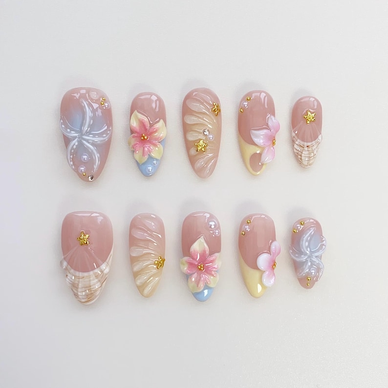 May include: A set of ten artificial nails in various designs. Each nail is a light pink color with detailed floral and abstract designs. Some nails feature 3D flowers in pink, yellow, and blue, while others have gold accents and pearl embellishments.