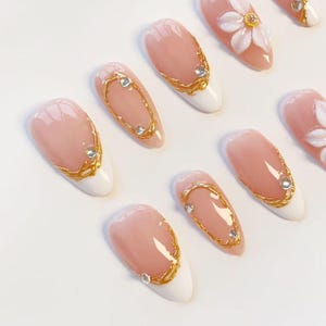 Puede incluir: Un juego de u&ntilde;as postizas en forma de almendra con base color nude y puntas blancas. Los dise&ntilde;os incluyen detalles de filigrana met&aacute;lica dorada, peque&ntilde;os diamantes de imitaci&oacute;n transparentes y un adorno floral blanco en relieve.