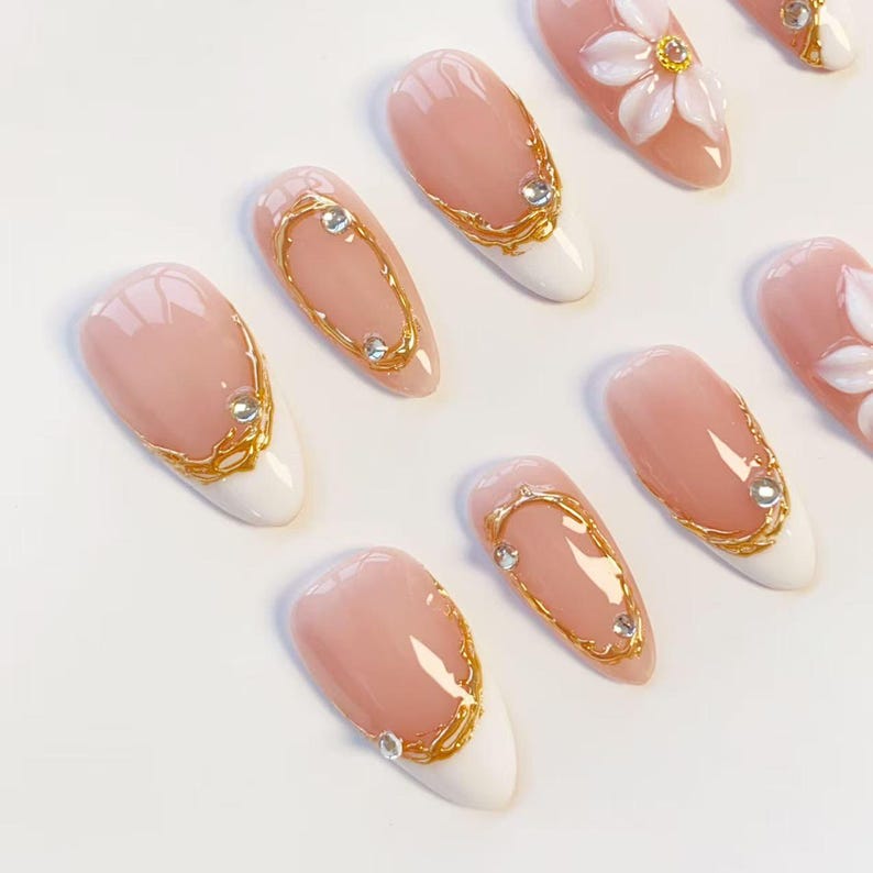 Puede incluir: Un juego de u&ntilde;as postizas en forma de almendra con base color nude y puntas blancas. Los dise&ntilde;os incluyen detalles de filigrana met&aacute;lica dorada, peque&ntilde;os diamantes de imitaci&oacute;n transparentes y un adorno floral blanco en relieve.