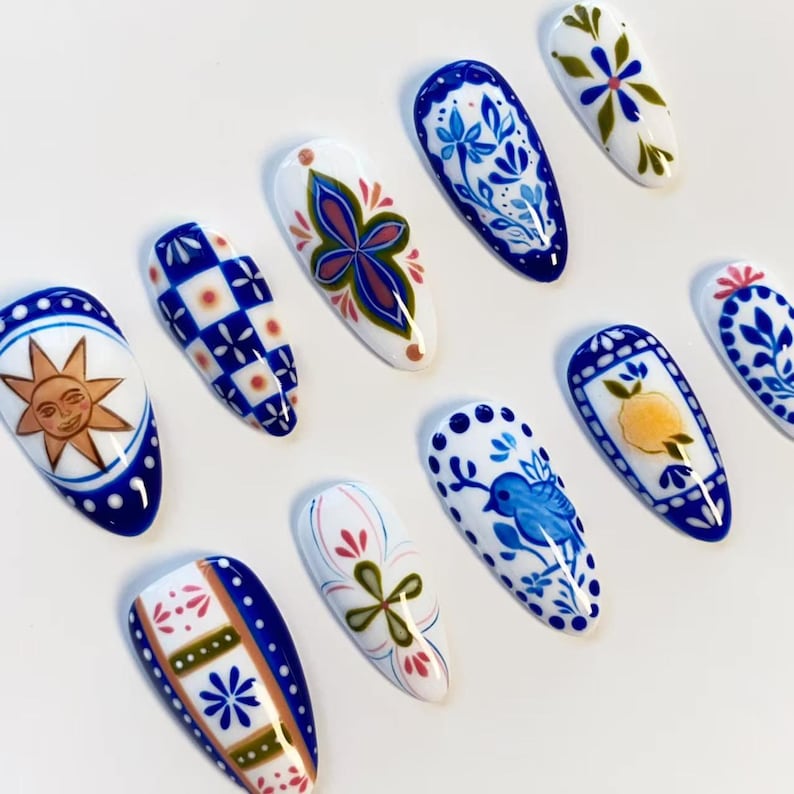 May include: A set of ten almond-shaped press-on nails featuring various hand-painted designs in blue, white, yellow, and green. Patterns include a sun face, blue and white checkered squares, floral motifs, a small blue bird, and a yellow lemon.