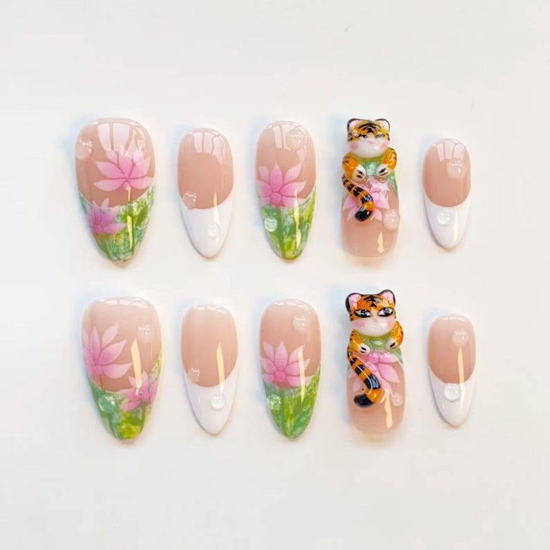 May include: A set of ten artificial nails. The nails feature a variety of designs, including pink lotus flowers, green leaves, and a 3D tiger figurine. Some nails have a white tip and a water droplet design.