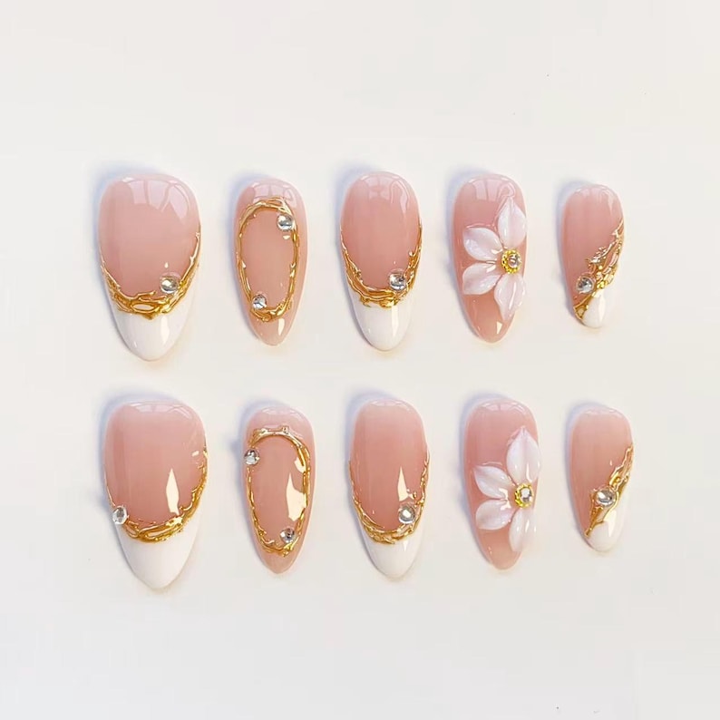 Puede incluir: Un juego de diez u&ntilde;as postizas en forma de almendra con base color nude y puntas blancas. Los dise&ntilde;os incluyen detalles dorados texturizados, peque&ntilde;os diamantes de imitaci&oacute;n transparentes y p&eacute;talos de flores blancos en relieve.