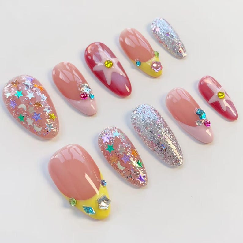 Pu&ograve; includere: Una collezione di dieci unghie finte in vari design. Alcune unghie presentano glitter, dettagli di stelle e lune e strass. Altre hanno un design a punta francese con accenti rosa e gialli. Le unghie sono di forma ovale e disposte su una superficie bianca.