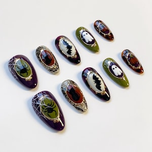 May include: A collection of Halloween-themed press-on nails. The nails feature designs such as spiders, bats, ghosts, and pumpkins. Colors include purple, green, orange, and black, with gold accents. The nails are oval-shaped and have a glossy finish.