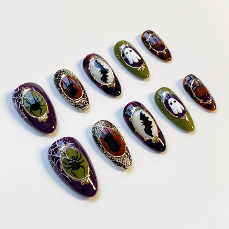 May include: A collection of Halloween-themed press-on nails. The nails feature designs such as spiders, bats, ghosts, and pumpkins. Colors include purple, green, orange, and black, with gold accents. The nails are oval-shaped and have a glossy finish.