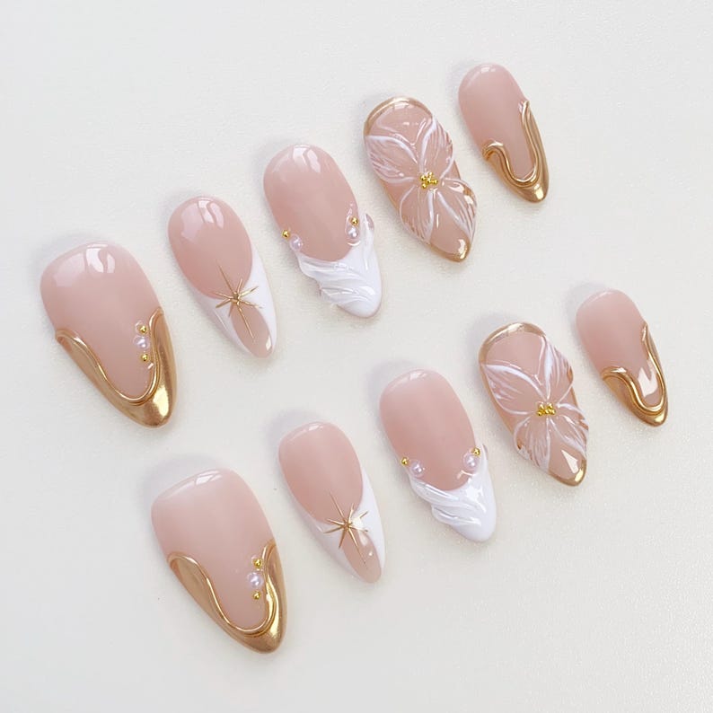 May include: A collection of almond-shaped false nails in a range of designs. The nails have a pale pink base with gold detailing, white floral and wave patterns, and starburst features. Some nails are adorned with tiny pearls.