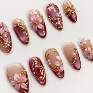 May include: A collection of artificial nails in various designs. Some nails are a light pink with gold accents and small pearls. Others are a deep red with gold trim and pearls. Several nails feature 3D pink flower designs.