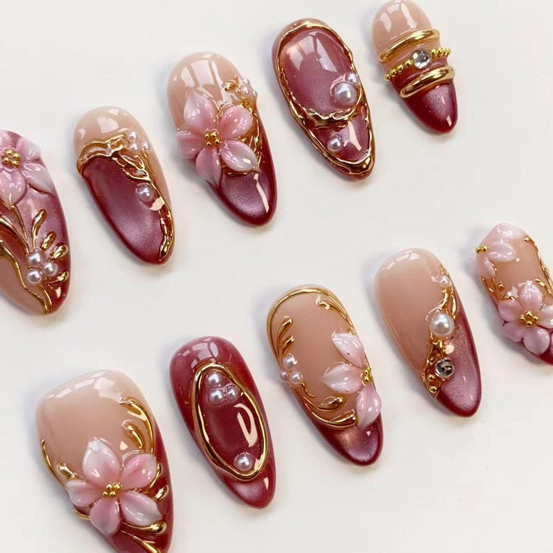 May include: A collection of artificial nails in various designs. Some nails are a light pink with gold accents and small pearls. Others are a deep red with gold trim and pearls. Several nails feature 3D pink flower designs.
