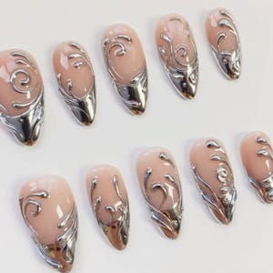 May include: Set of ten artificial nails with a light pink base and silver metallic accents. Each nail features a silver swirl design and a silver tip, creating a glamorous and elegant look. The nails are arranged on a white surface.