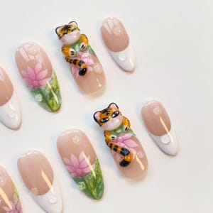 May include: A set of artificial nails with a variety of designs. Some nails feature a pink and white French tip, while others have a pink lotus flower design with green leaves. Two nails have a 3D tiger figurine.