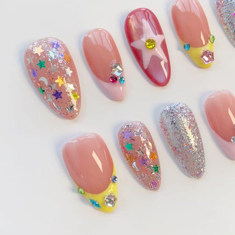 Pu&ograve; includere: Una collezione di unghie finte con vari design. Alcune unghie presentano glitter, paillettes a forma di stella e strass. Altre hanno un design a french manicure rosa e giallo. Un'unghia ha un disegno a stella con una gemma gialla.