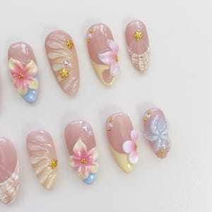 May include: A set of almond-shaped artificial nails with a variety of floral and abstract designs. The nails feature a base of pink and nude tones, with accents of yellow, blue, and gold. Some nails have 3D flower embellishments.