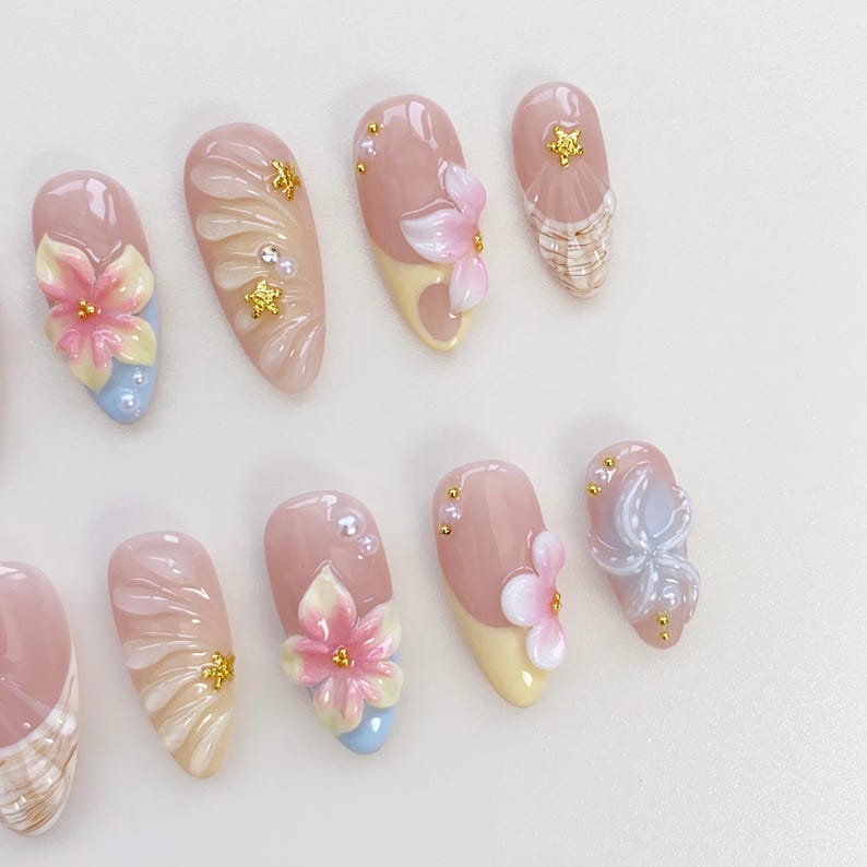 May include: A set of almond-shaped artificial nails with a variety of floral and abstract designs. The nails feature a base of pink and nude tones, with accents of yellow, blue, and gold. Some nails have 3D flower embellishments.