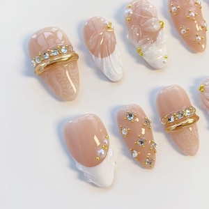 May include: A collection of artificial nails in various designs. Some nails are nude-colored with gold and rhinestone accents, while others feature white tips and pearl or floral embellishments. The nails showcase intricate lace patterns and a variety of decorative elements.